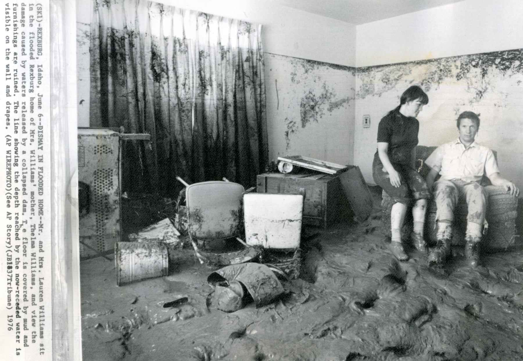 Mr. and Mrs. Lauren Williams sit in the flooded Rexburg home of Mrs. Williams' mother, Thelma Williams, and view the damage caused by waters released by a collapsed dam. The floor is covered by mud and furnishings are ruined. The line showing the depth reached by the now-receded water is visible on the wall and drapes (photo: AP Wire / waterarchives.org). flooded rexburg home teton dam flood